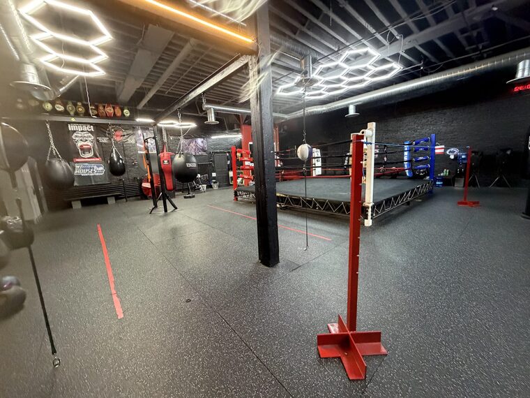 A row of heavy bags hanging along the gym floor, with the ring and its blue corner posts beyond them.