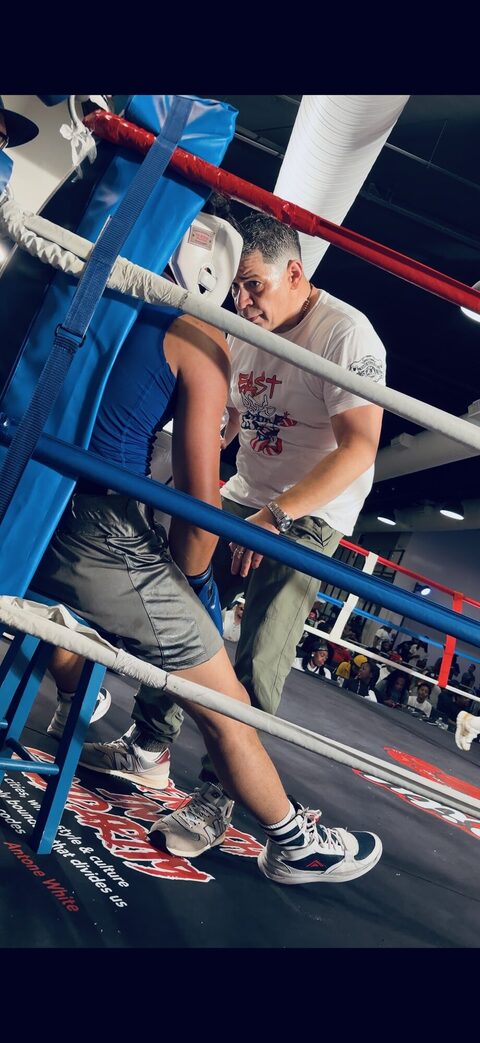 A coach leaning over the blue corner ropes to talk to his fighter between rounds, the arena crowd behind them.