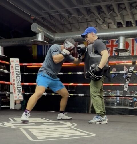 A boxer in blue shorts throwing a hook into a coach's focus mitts inside the ring.
