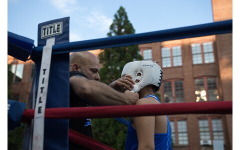 A coach fitting headgear onto a young boxer at the ring apron before his bout.